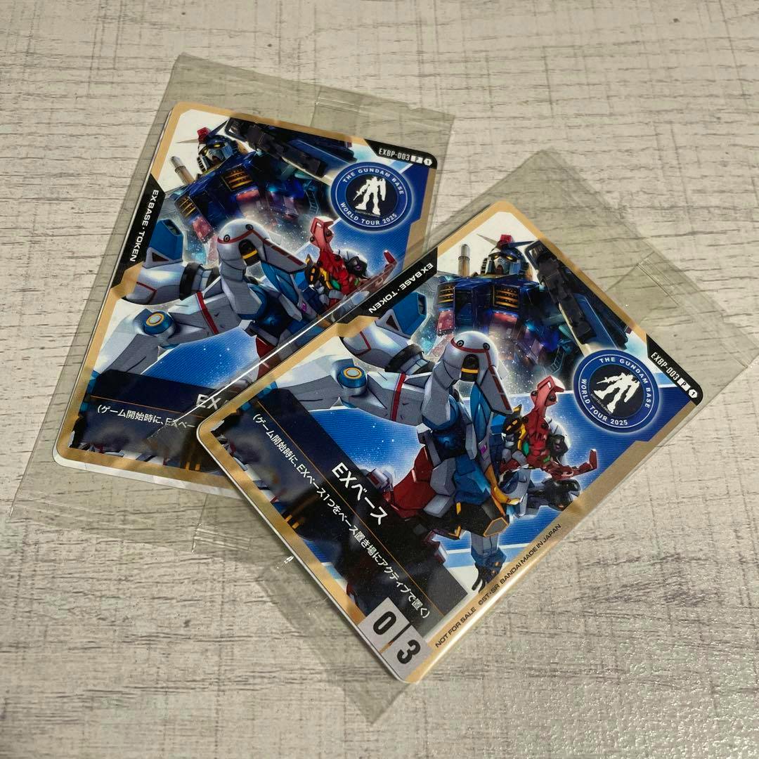 Two Gundam Base EXベース photocard tokens in clear sleeves placed on a light wood surface.