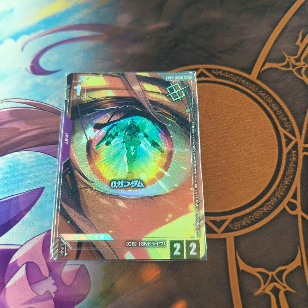 A sleeved Gundam Card Game trading card front showing artwork of an eye reflecting a Gundam and the text "0 ガンダム" placed on a playmat.