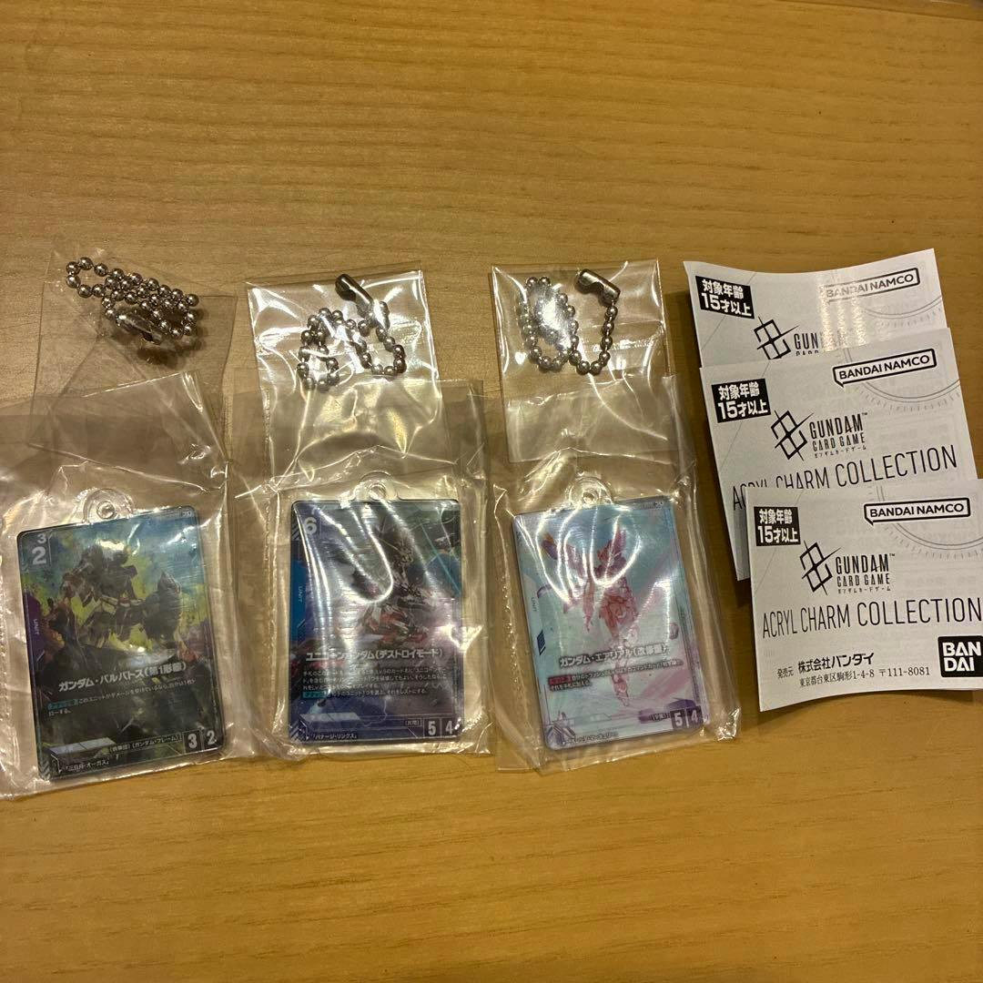 Photo of three Gundam Card Game acrylic charm keychains sealed in plastic with their ball chains and product papers on a wooden surface.