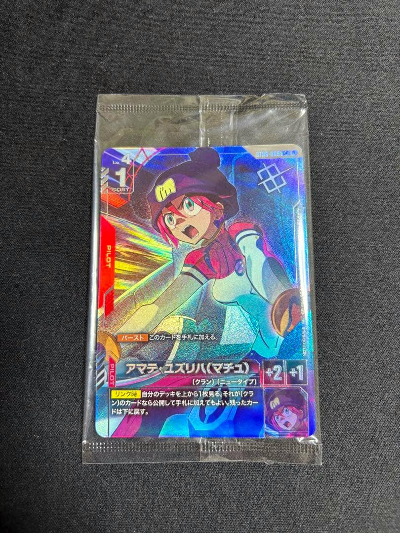 Front of the sealed Gundam trading card アマテ・ユズリハ(マチュ) showing holographic character art in a clear plastic sleeve on a black background.