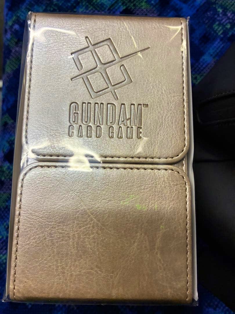 A metallic card case with the embossed text "GUNDAM CARD GAME" on the front, shown in its plastic packaging.