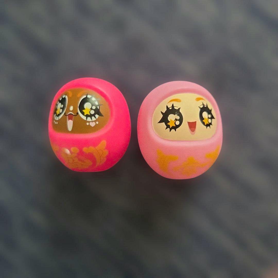 Two pink capsule-toy daruma-style figures with star-shaped pupils on a gray background.
