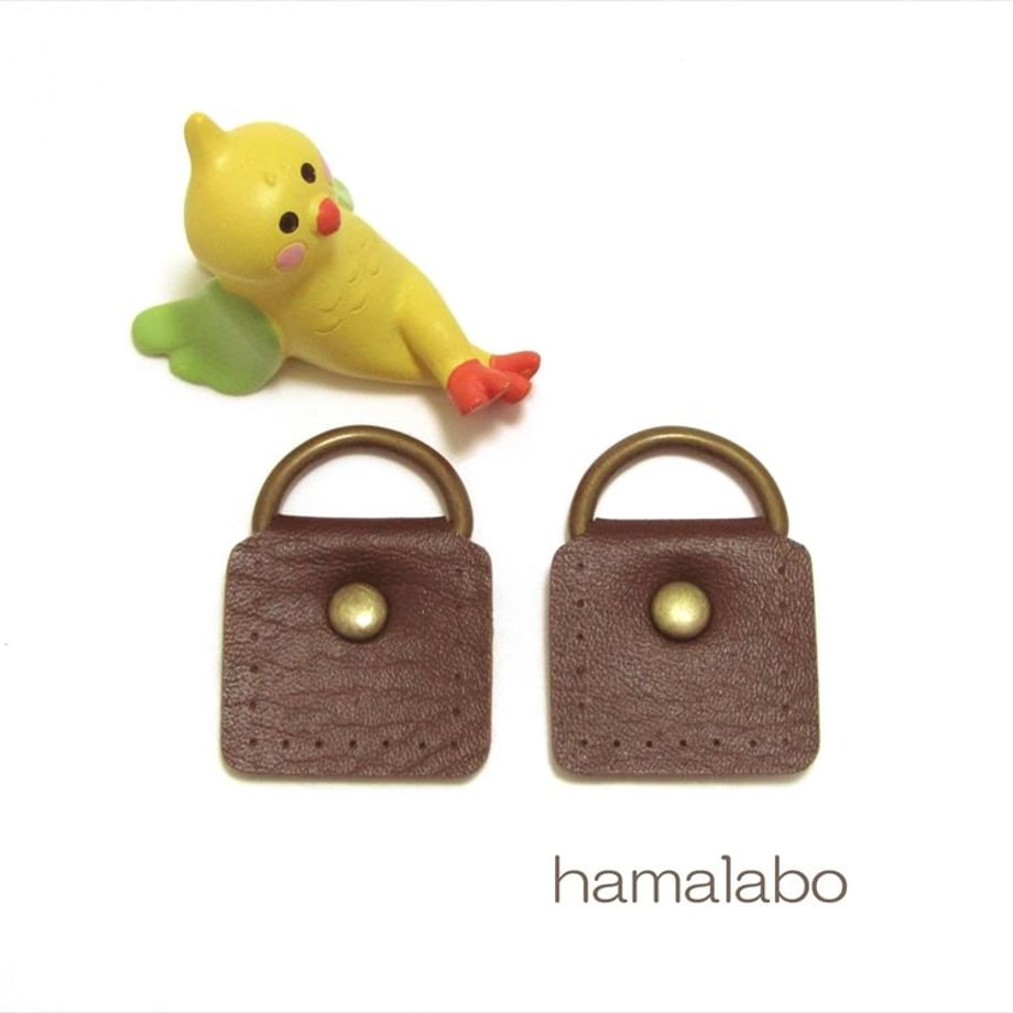 Two small brown cow-leather shoulder-bag attachments with brass D-rings shown on a white background next to a small yellow bird figurine.