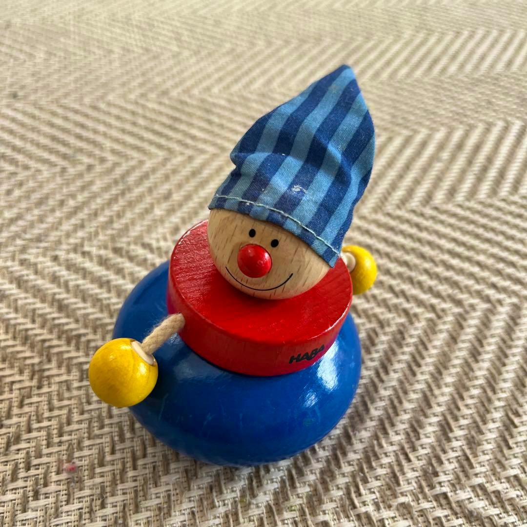 Wooden HABA rattle toy with a blue base, red ring, smiling wooden head wearing a blue striped hat and yellow bead hands on a woven surface.