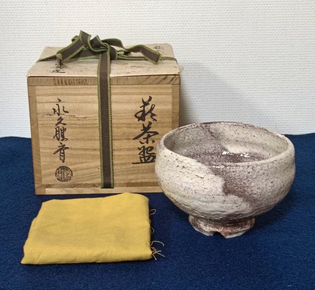 A Hagi tea bowl displayed beside its wooden box and a yellow cloth on a blue surface.