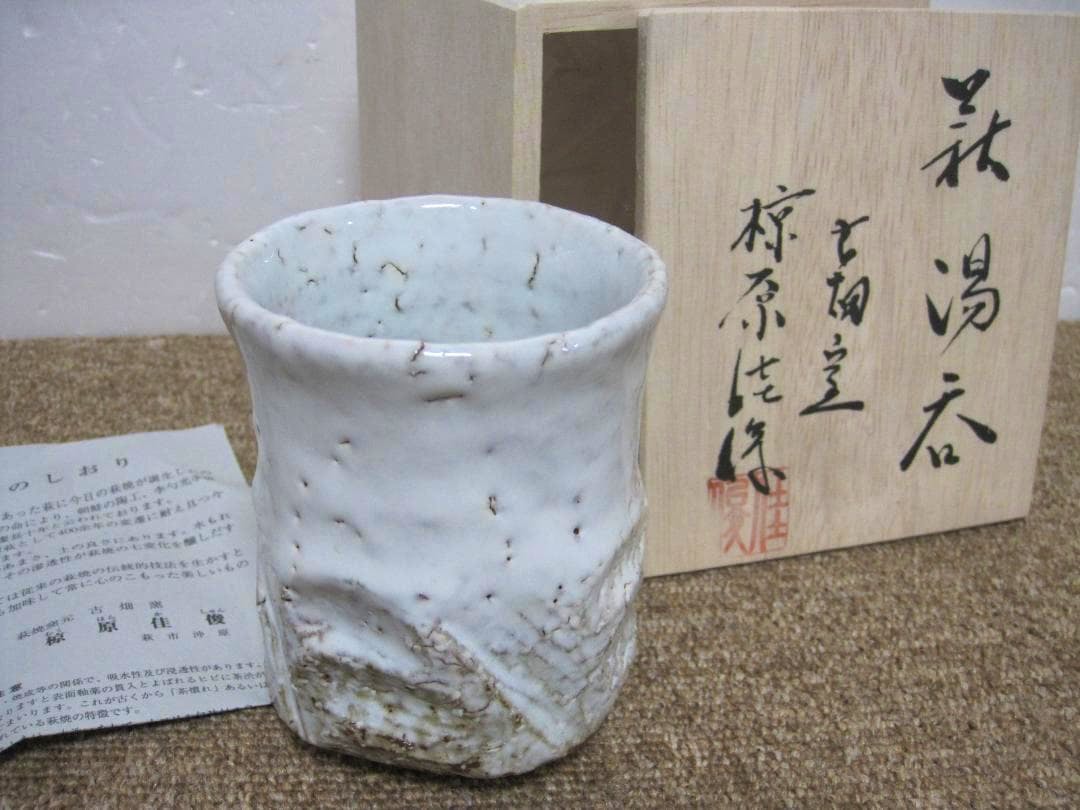 White-glazed Hagi-yaki yunomi (tea cup) shown with its wooden box and a paper certificate in the background.