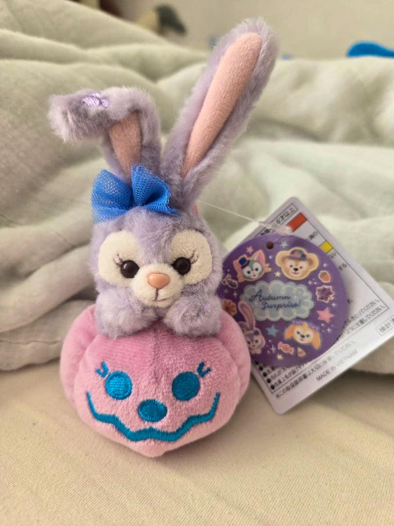 A purple Stella Lou plush charm sitting on a pink jack-o'-lantern plush with its retail tag visible.