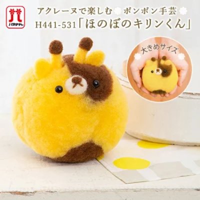 A yellow and brown felted round giraffe mascot pompom made of wool shown on a table with an inset of hands holding it.