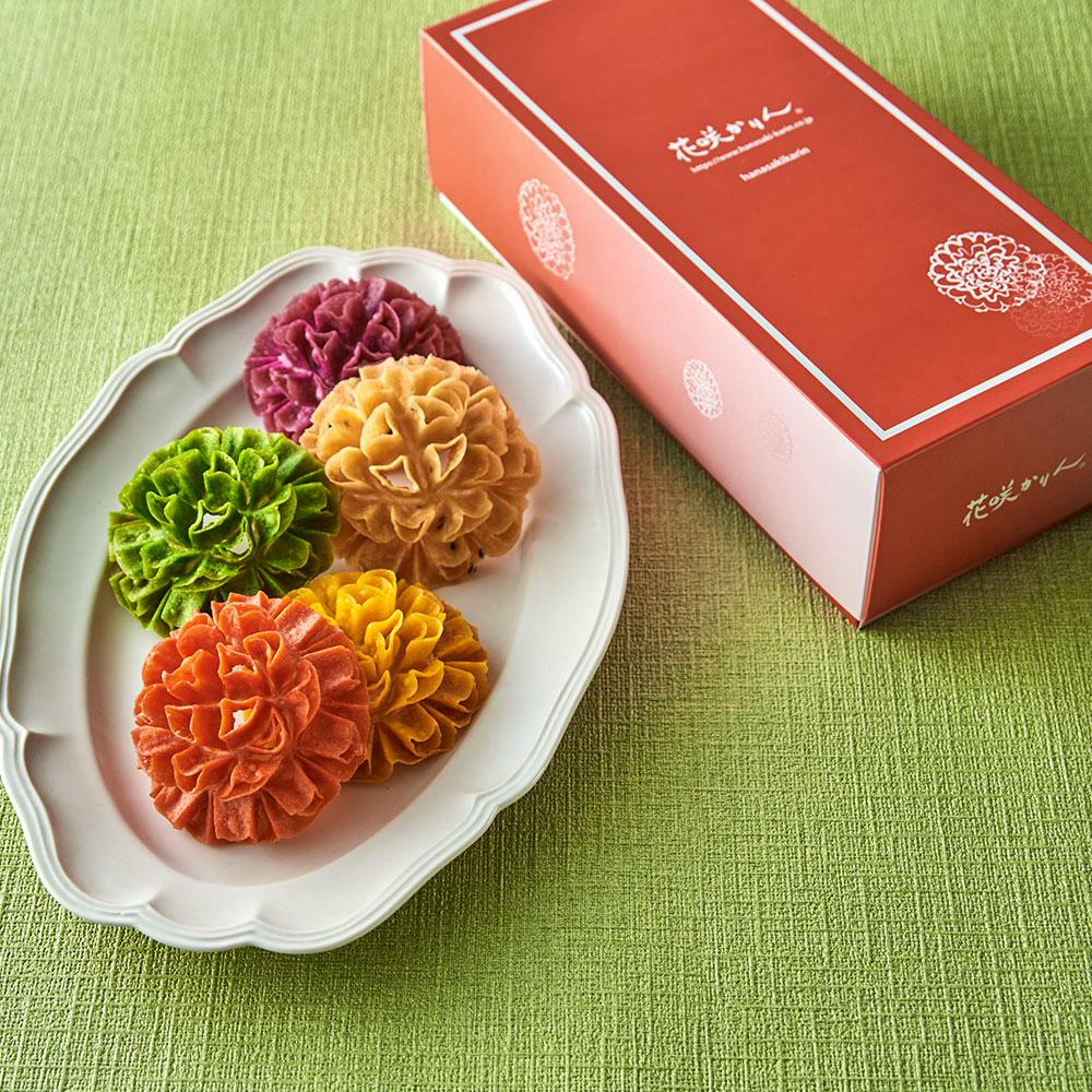 A white plate holding five colorful flower-shaped rice crackers next to a red product box on a green tablecloth.