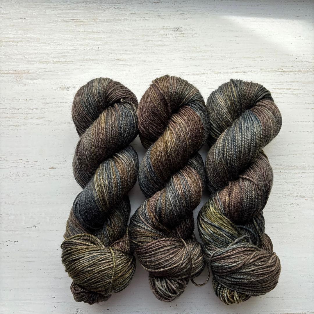 Three hand-dyed sock yarn skeins (潮風とオリーブ) in brown, blue, and green tones arranged on a white wooden surface.