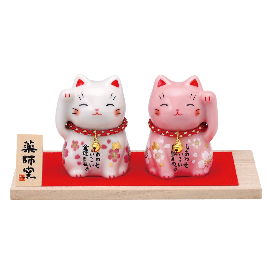 Two painted ceramic maneki-neko (beckoning cat) figures with cherry blossom decoration displayed on a wooden base with red felt.