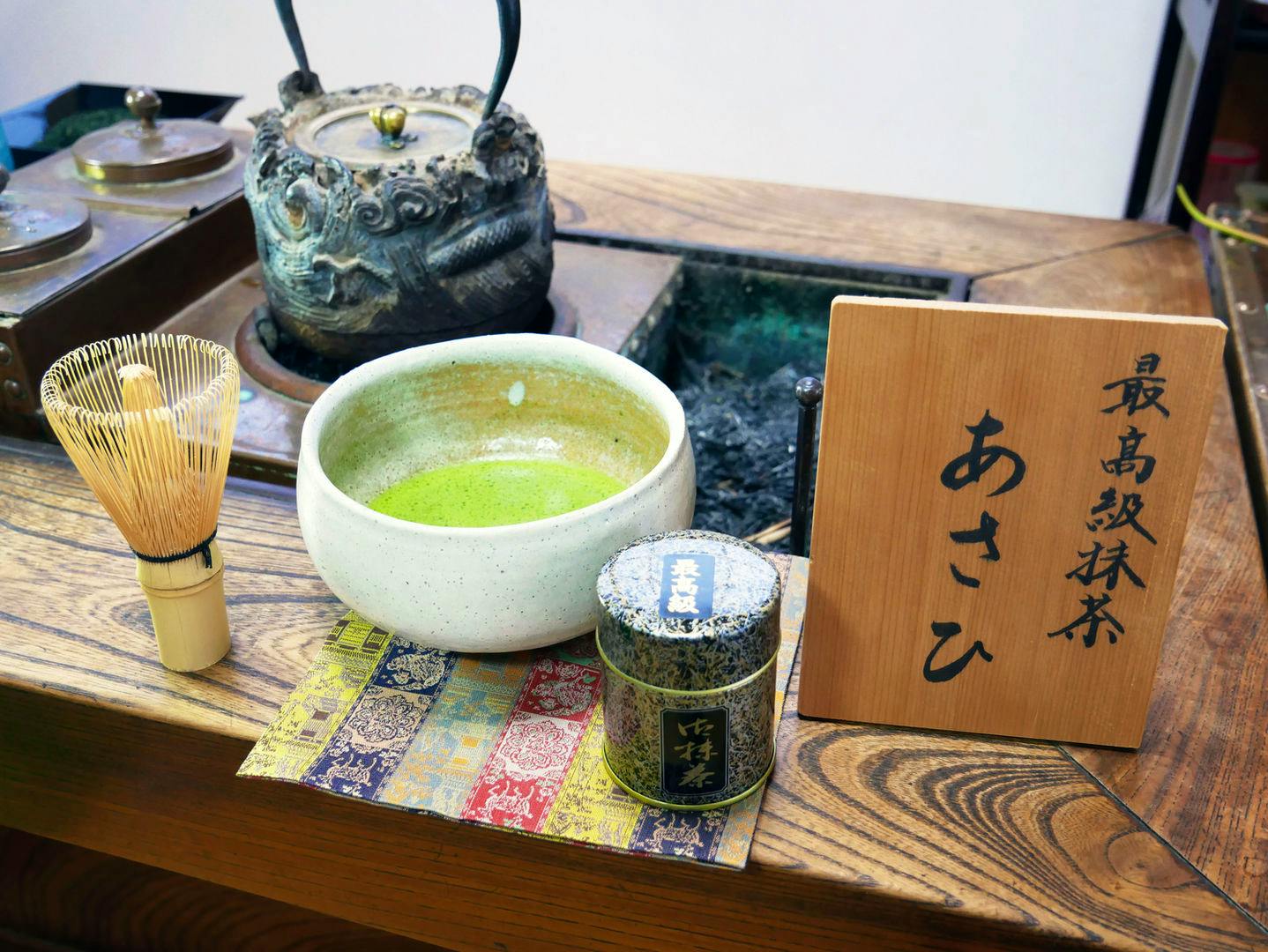 A ceramic bowl of prepared matcha, a bamboo whisk, and a small matcha tin displayed on a wooden table next to a wooden sign.