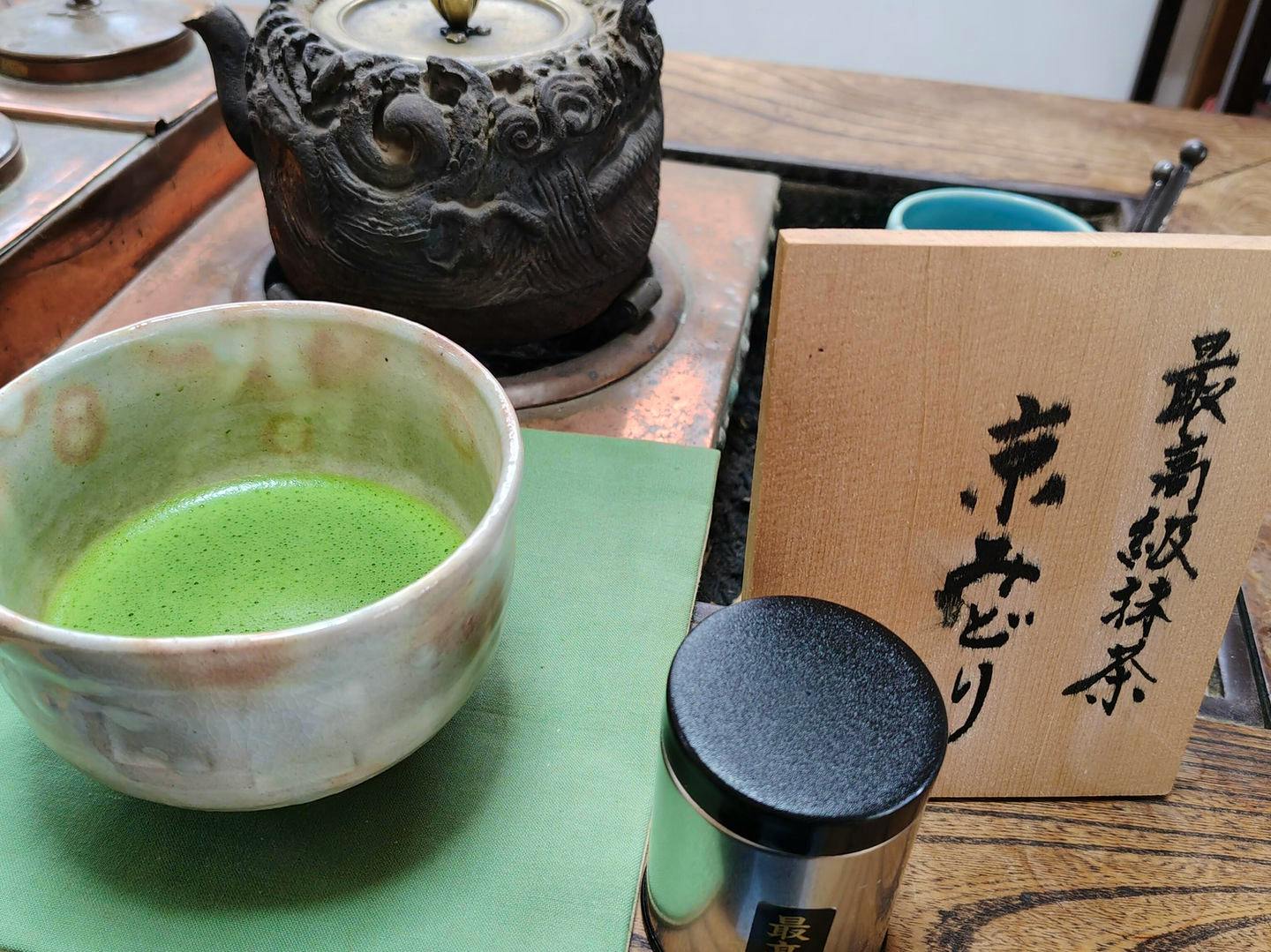 A bowl of prepared matcha next to a small metal tea canister and a wooden board with Japanese writing.