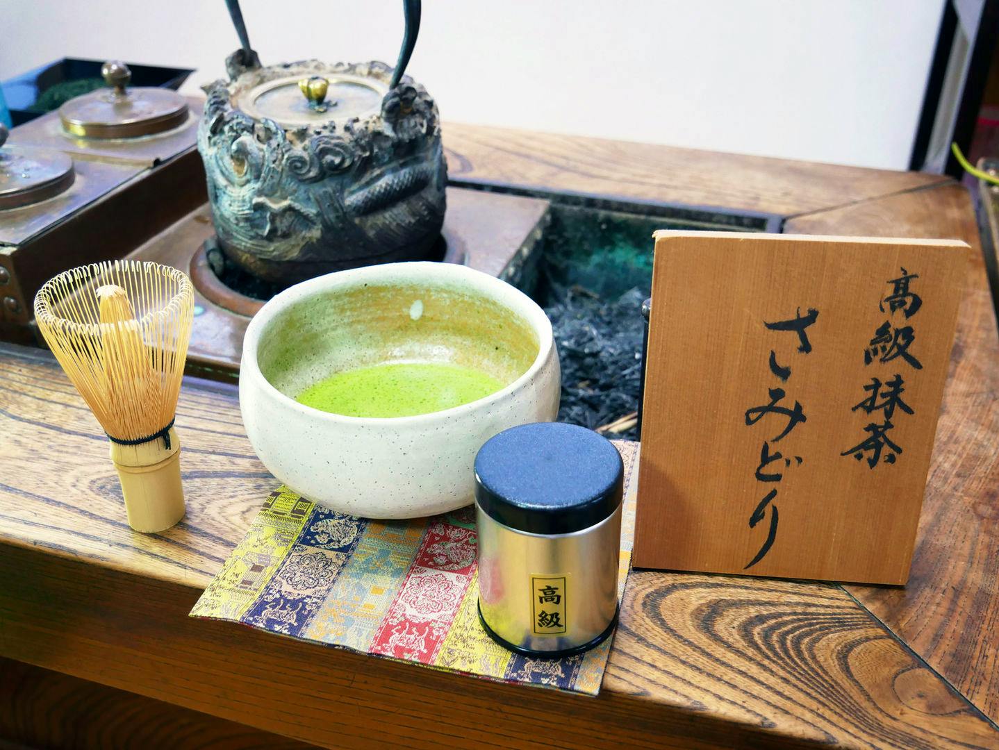 A tea bowl of prepared matcha, a bamboo whisk, a small silver tea can, and a wooden sign with Japanese calligraphy on a wooden table.