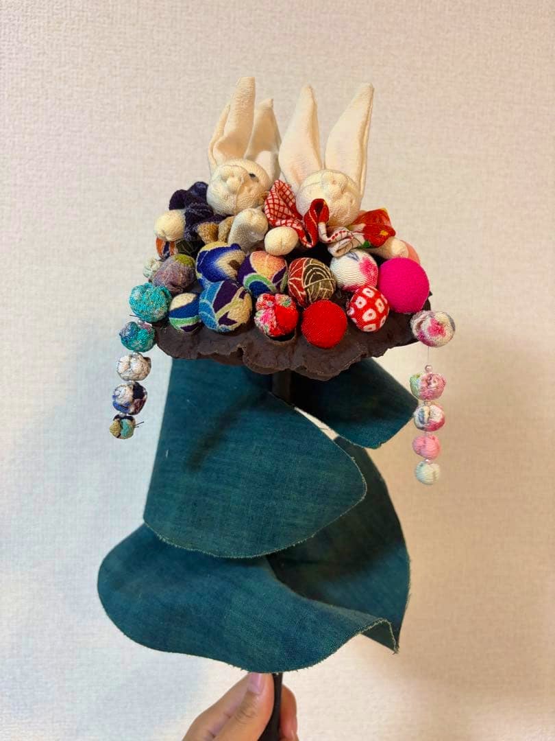 Handmade chirimen fabric lotus-seed decoration with two small fabric rabbit figures and colorful cloth balls mounted on a stem with green fabric leaves.