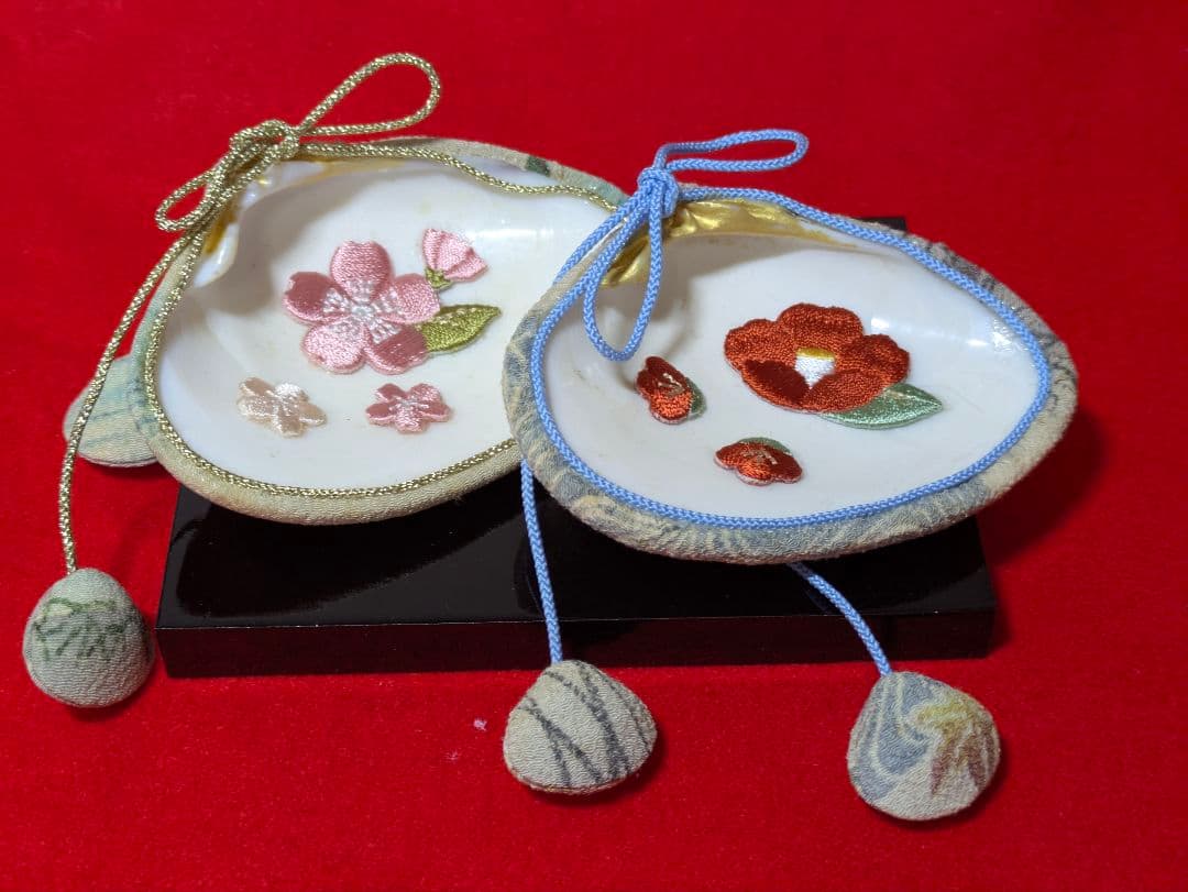 A pair of handmade chirimen fabric shell ornaments with embroidered flowers and hanging fabric balls on a red background.