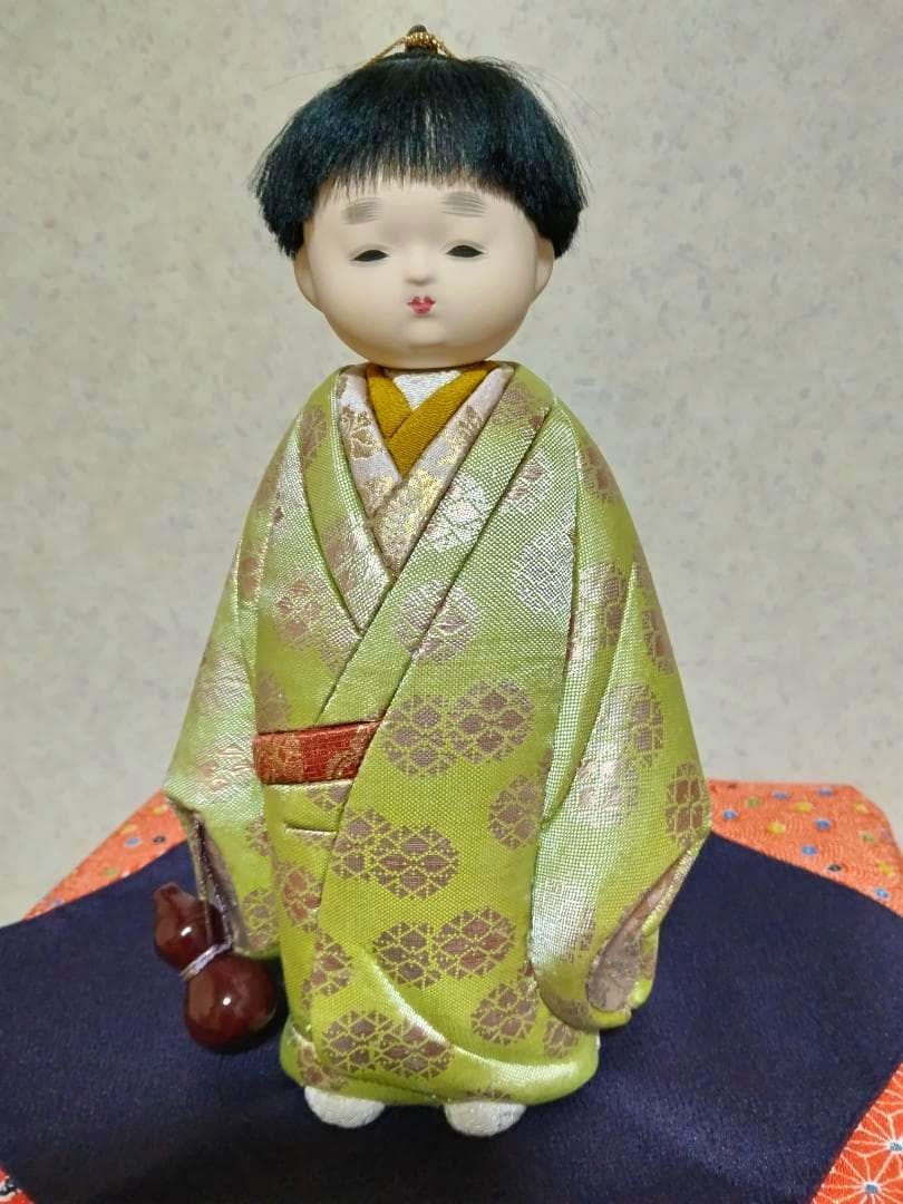 A plastic kimekomi doll wearing a green patterned kimono standing on a cloth cushion.