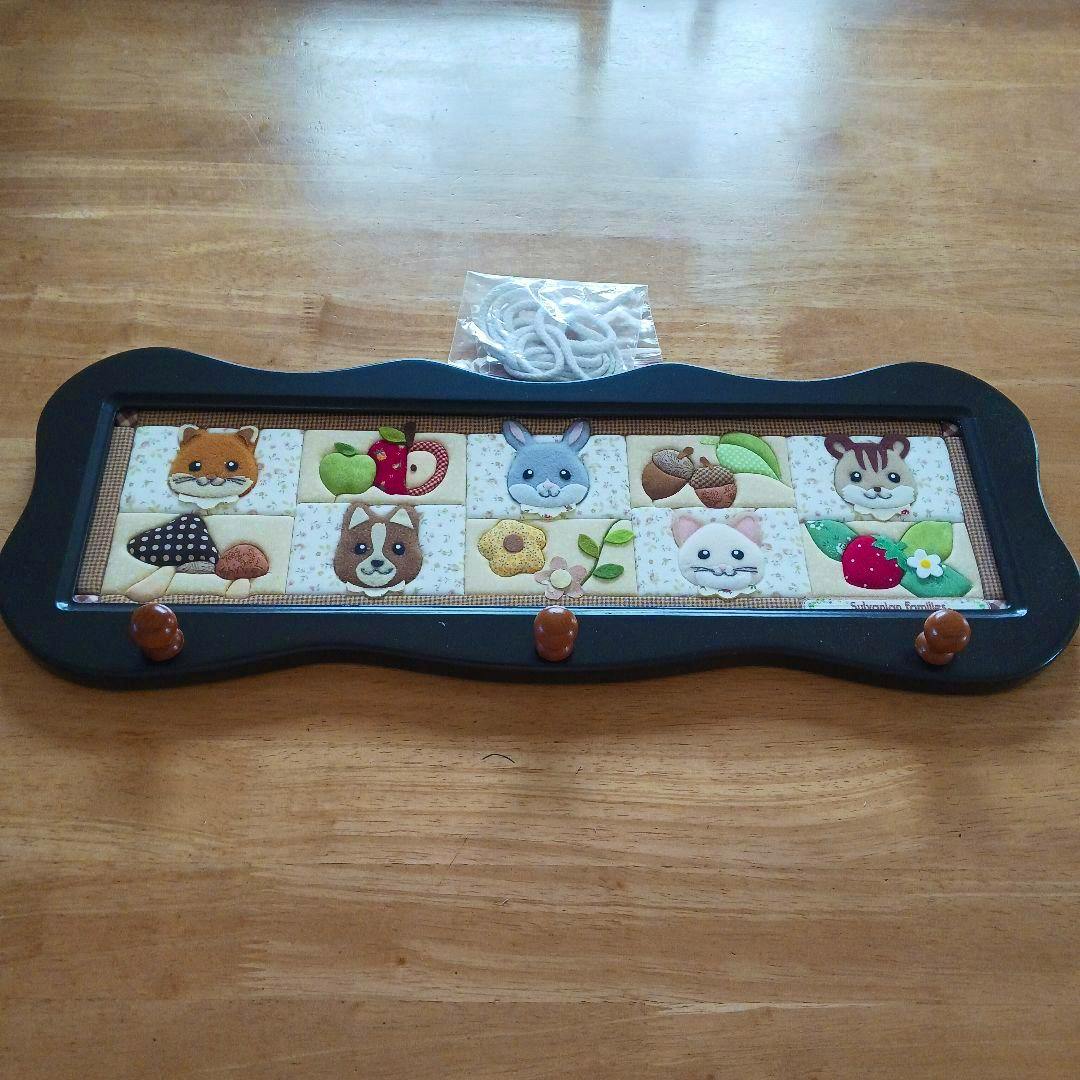 A rectangular wall hook with three brown pegs and a decorative patchwork panel of animal faces, shown on a wooden surface with a small coiled rope above.