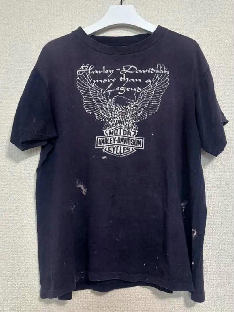Front view of a men's cotton Harley‑Davidson T-shirt with a white eagle and Harley‑Davidson logo print, hanging on a white hanger against a wall.