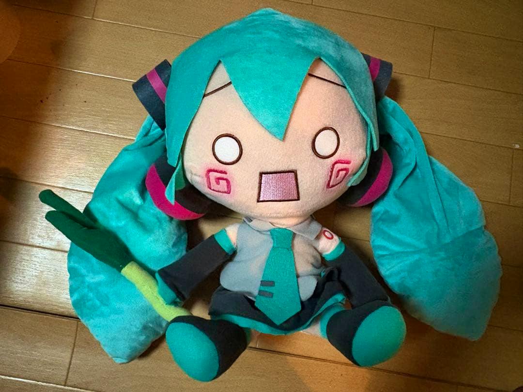 A Hachune Miku mega jumbo plush toy with teal twin-tails and an embroidered surprised face, sitting on a wooden floor and holding a green leek.