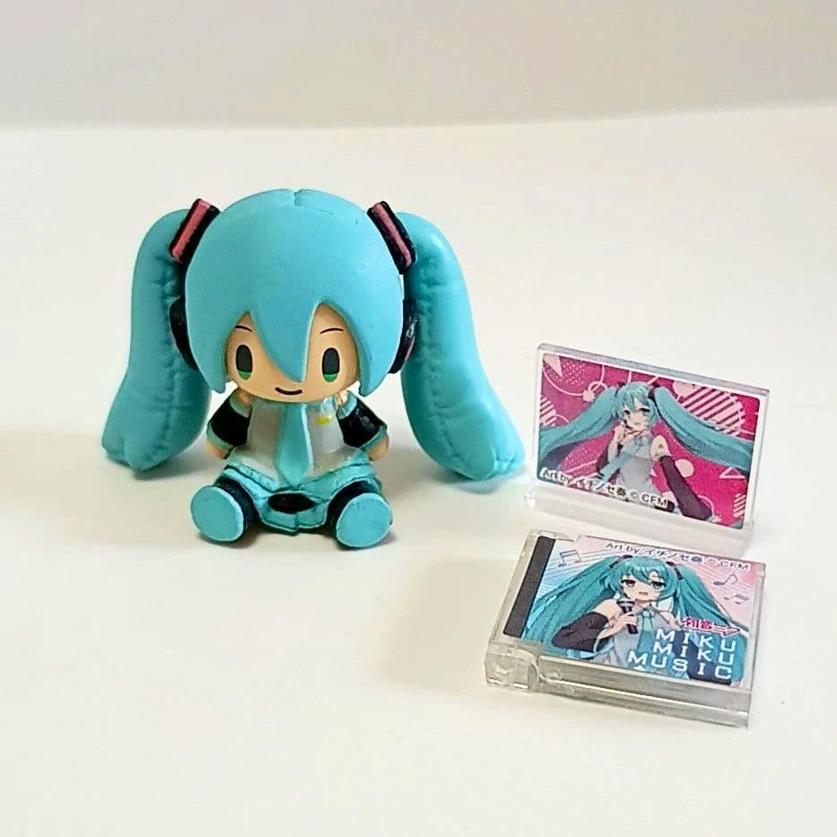 A small chibi Hatsune Miku plastic figure sitting on a white surface with two miniature CD/album cases beside it.