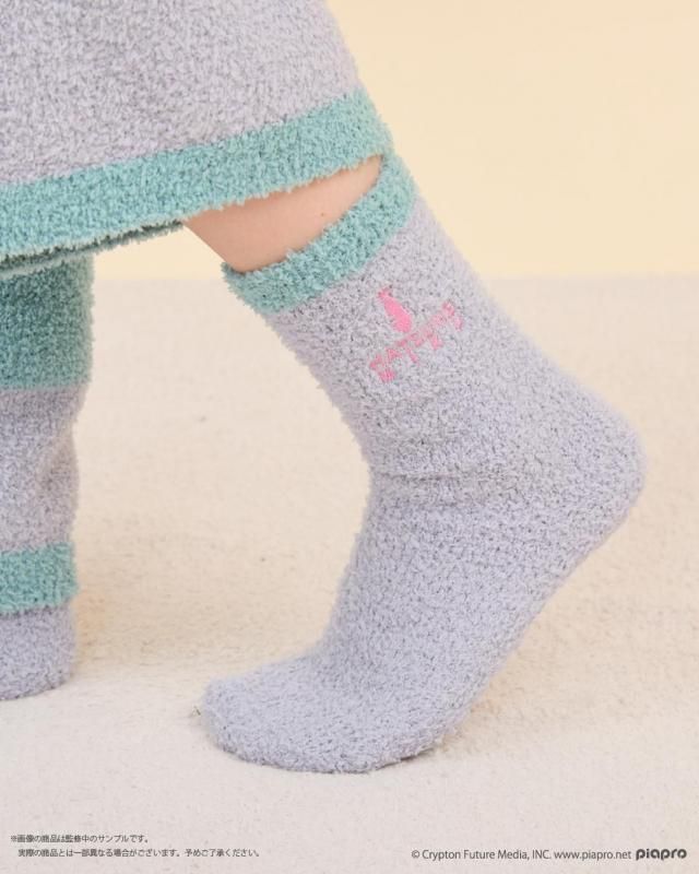 Fuzzy light-blue and mint-green room socks with a pink Hatsune Miku logo worn on a person's lower legs.