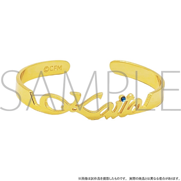 Gold-colored metal open name bangle reading "KAITO" with a small blue gem, shown on a white background.