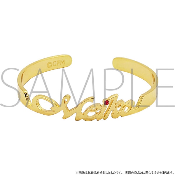 Gold metal name bangle spelling "Meiko" in cursive with a small red stone, photographed on a white background.