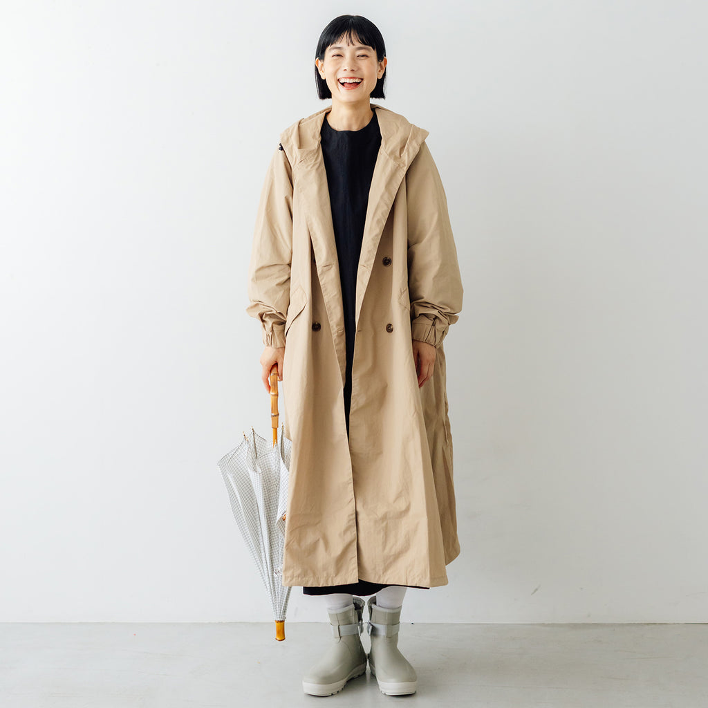 Model wearing a beige HAU spring & raincoat in a front view, standing and holding a folded umbrella.