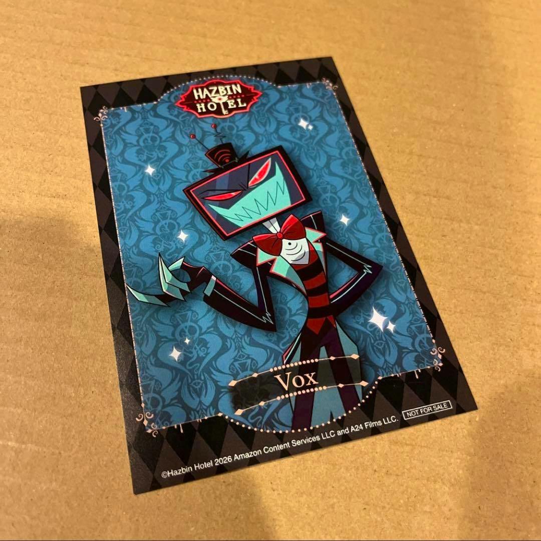 Photograph of a Hazbin Hotel postcard featuring the character Vox on a blue patterned background, lying on a brown surface.