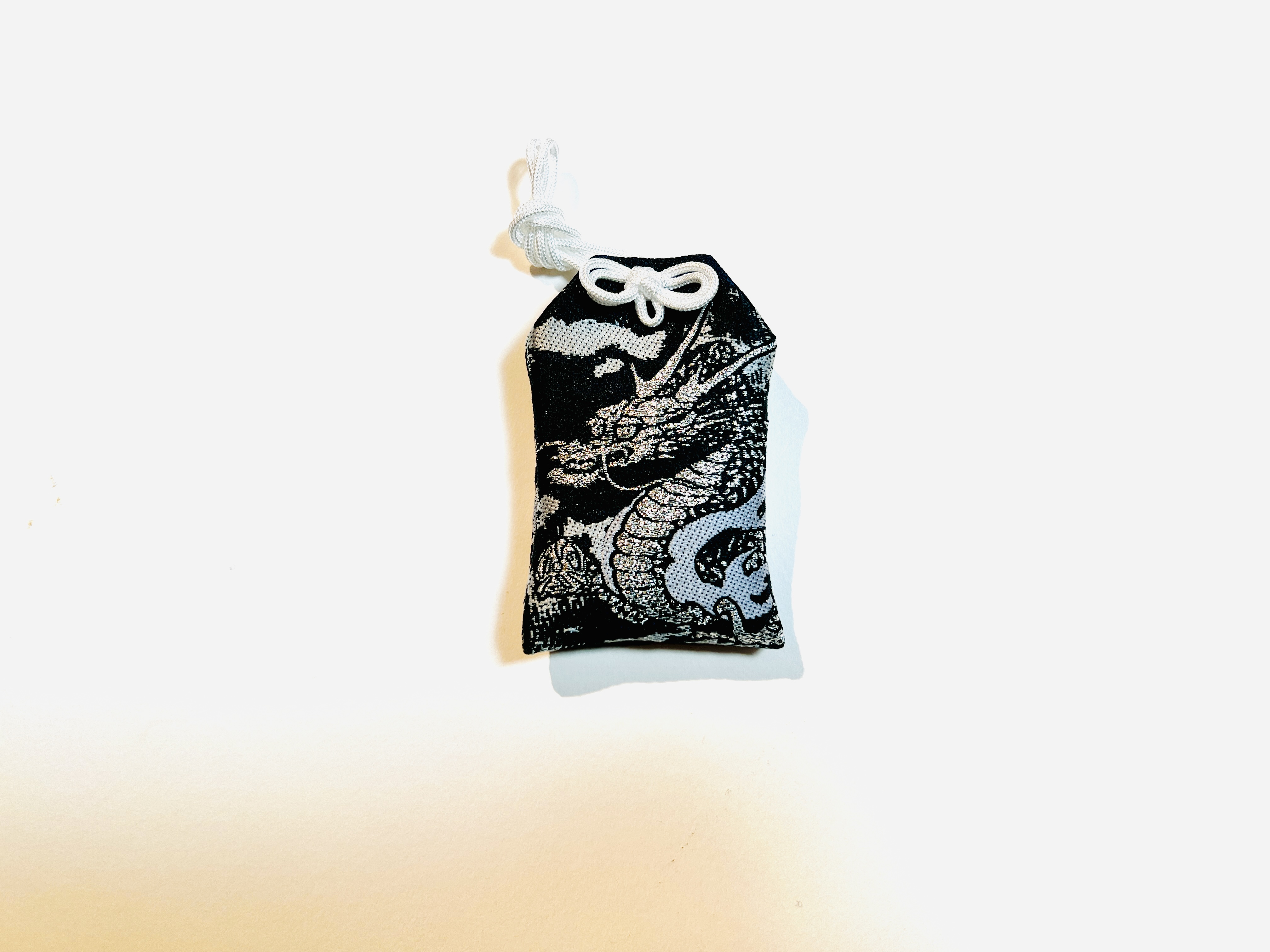A black cloth amulet with a silver dragon pattern and white cord on a white background.