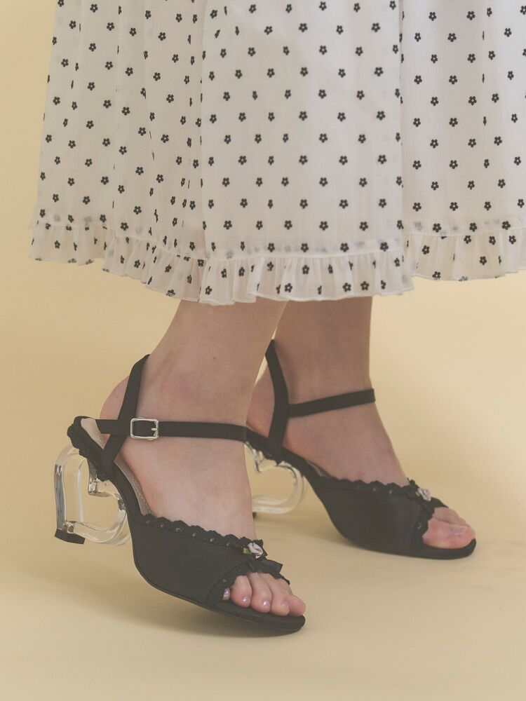 Black strap sandals with clear heart-shaped heels worn on a model's feet beneath a white skirt with a small floral print.