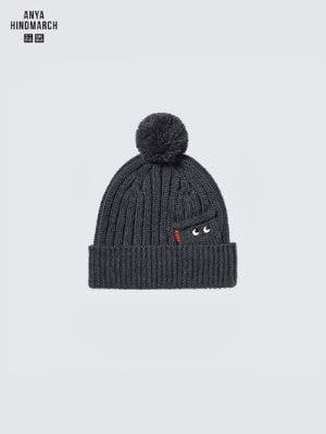 A grey knit beanie with a pom-pom and a small pocket showing embroidered eyes.
