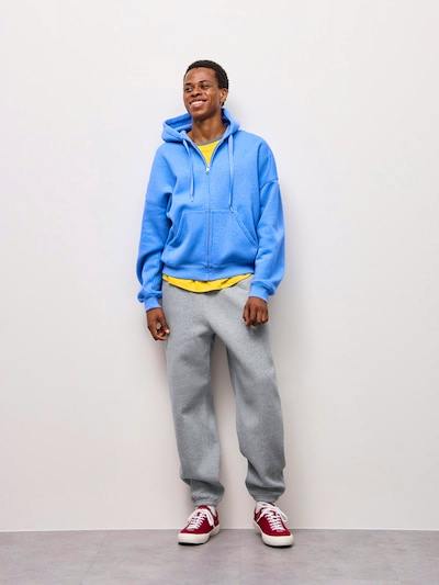 Male model standing against a light background wearing the grey sweat jogger pants with a blue hoodie and red sneakers.