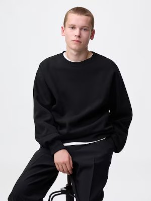 Person seated on a stool wearing a black sweatshirt and black trousers against a plain background.