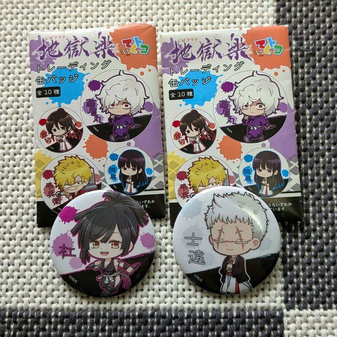 Two 地獄楽 trading badge boxes and two round can badges with chibi character artwork on a textured fabric background.