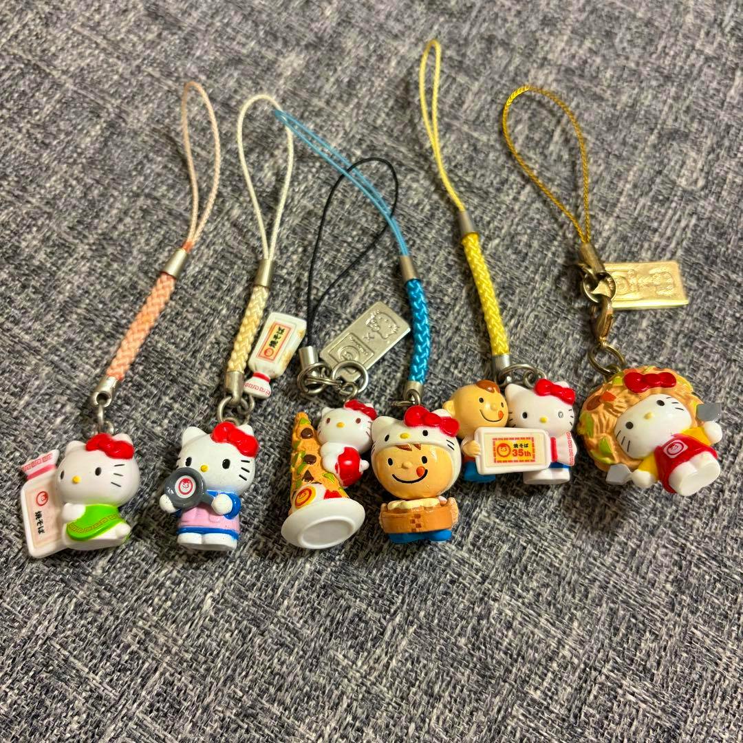 Six Hello Kitty Maruchan collaboration plastic keychain straps arranged in a row on a gray fabric background, showing the character designs.