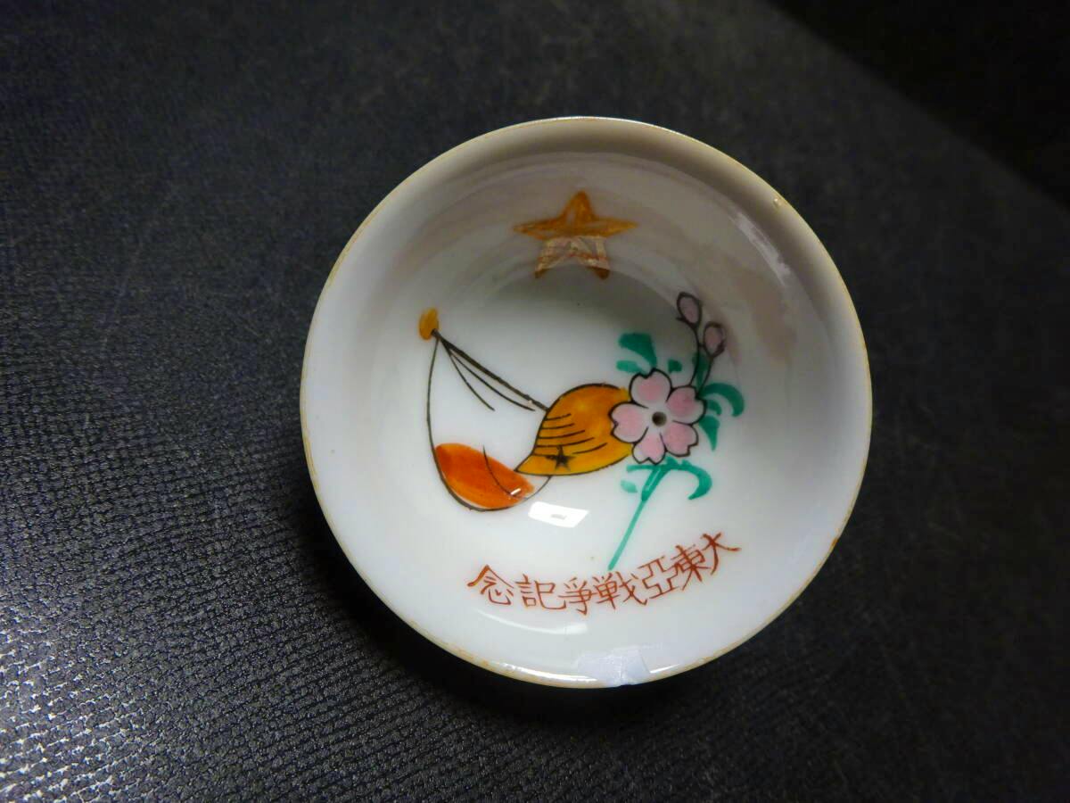 Top view of a small ceramic cup/plate showing a painted helmet, flower motif, and Japanese inscription.
