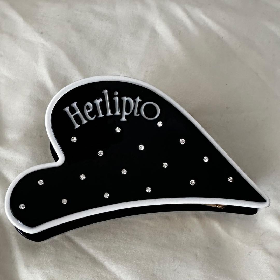 Black heart-shaped Herlipto hair clip with white trim and scattered rhinestones on a light fabric background.