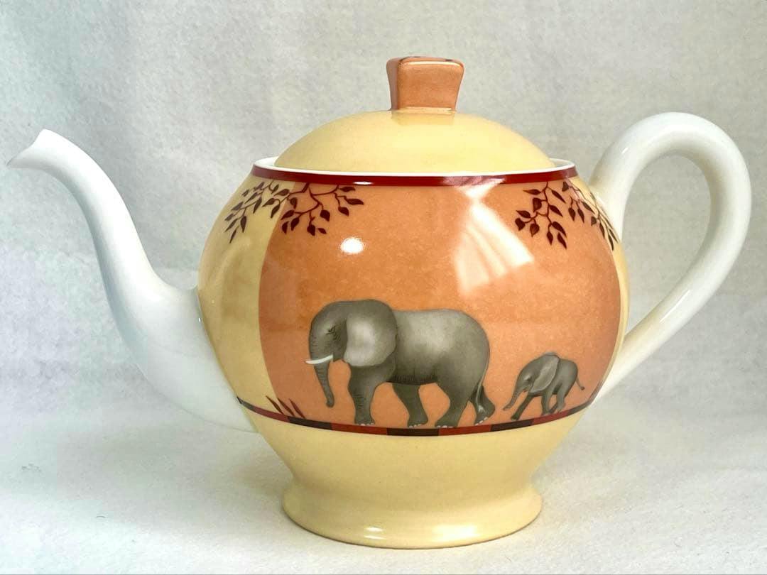Side view of the HERMES teapot showing its elephant decoration, lid, spout, and handle on a cream and orange background.
