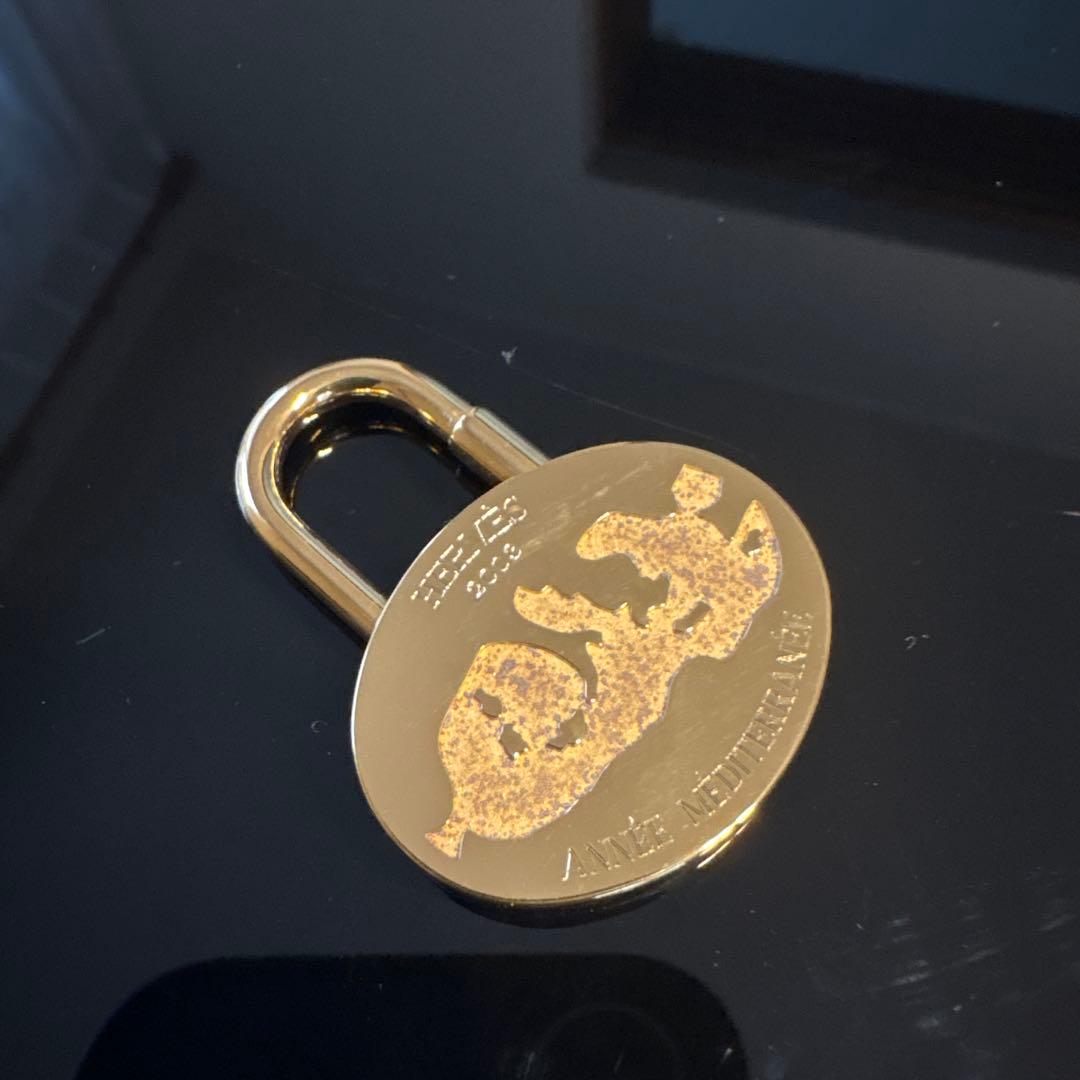 Hermes ANNEE MEDITERRANEE gold padlock with a decorative motif resting on a black reflective surface.