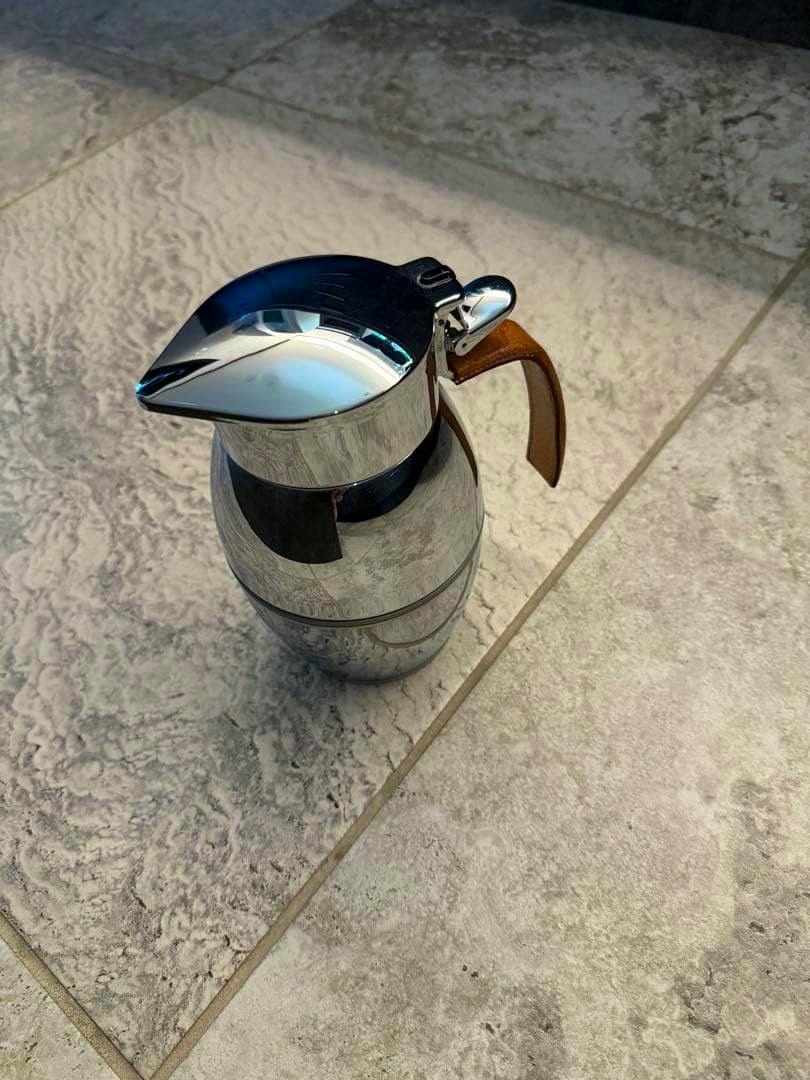 A stainless steel HERMES thermal pot with a brown leather handle on a tiled floor.