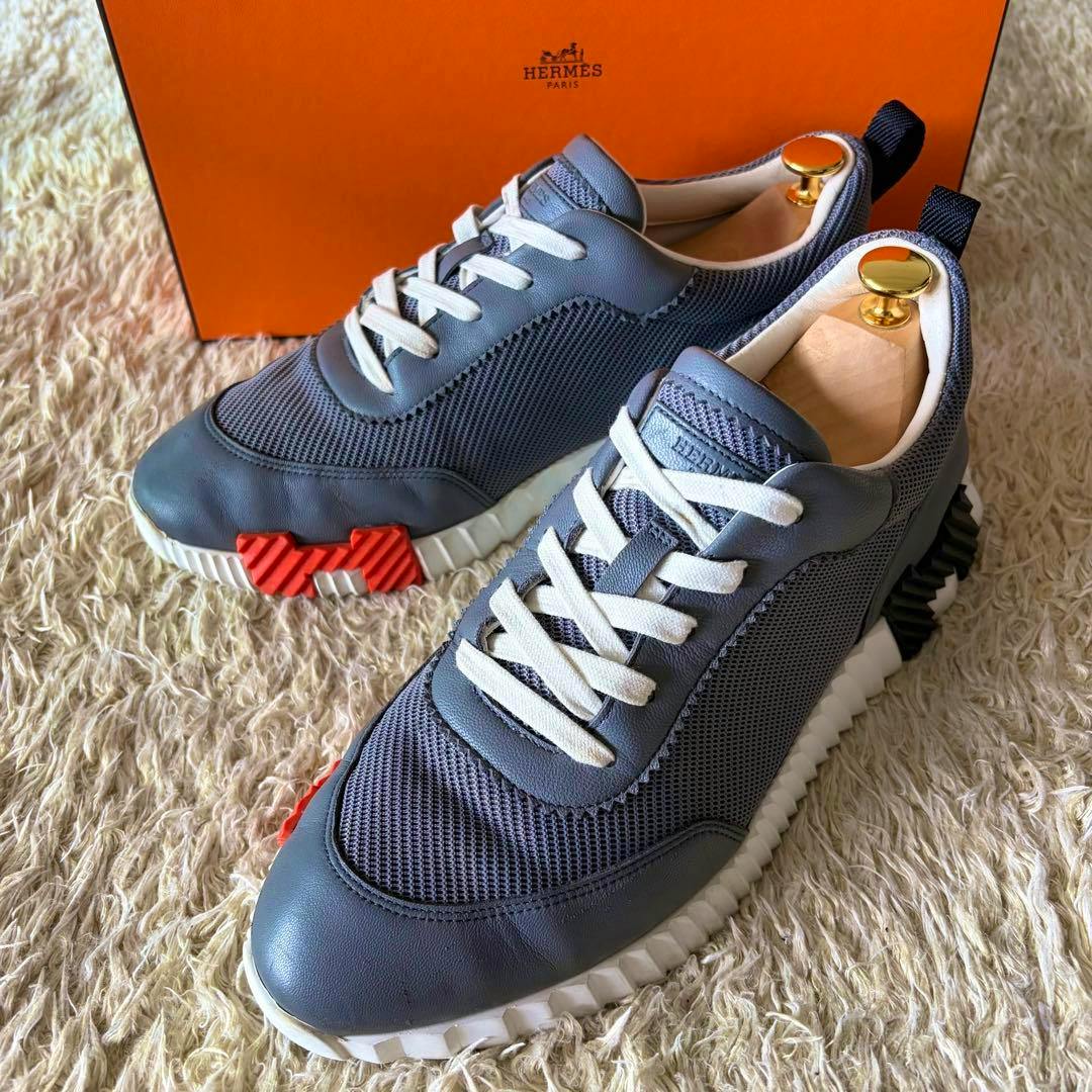 Pair of grey HERMES Bouncing leather sneakers (size 42) on a rug with an orange HERMES box behind.