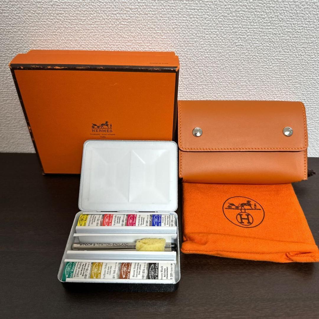 Open compact watercolor paint set shown with a metal paint tin, orange Hermès leather case, orange cloth pouch, and orange box on a dark surface.
