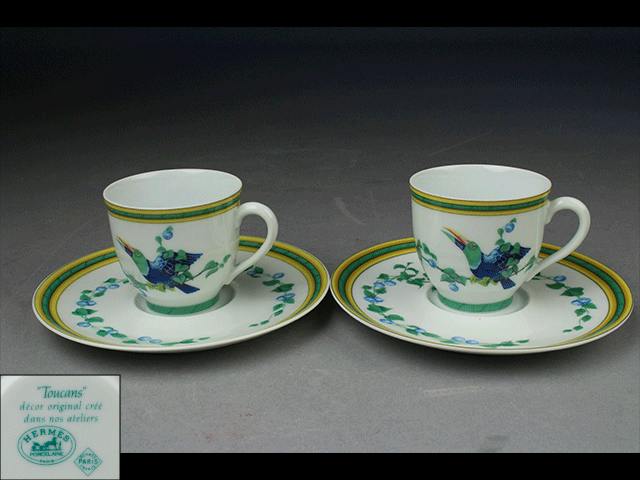Pair of HERMES porcelain cups and matching saucers with green-and-yellow rims and toucan decorations on a gray background.