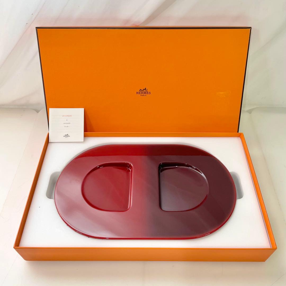 Red oval Hermès decorative tray displayed inside an open orange Hermès box with a small card.