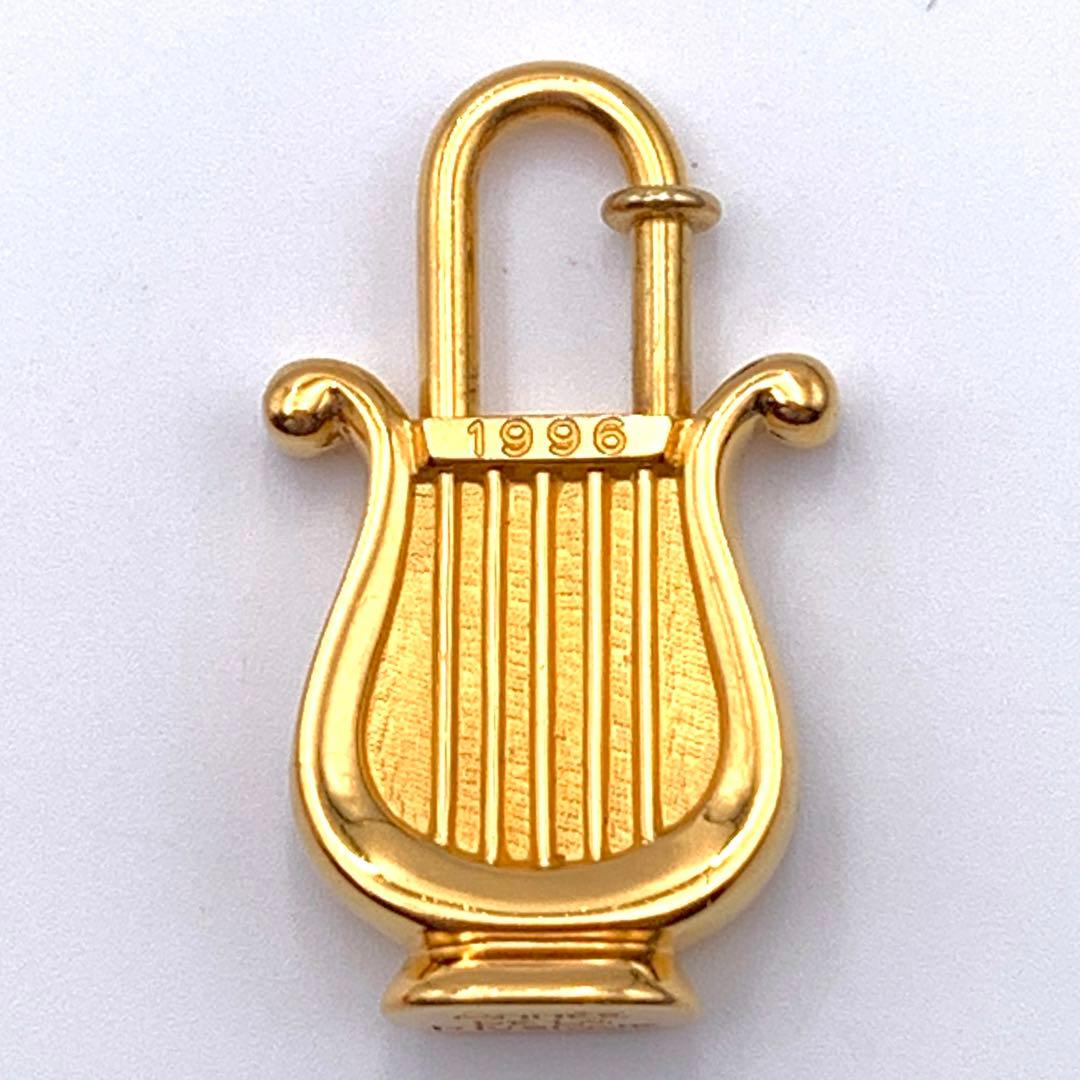 Front view of a gold harp-shaped Hermes padlock charm marked "1996" on a white background.