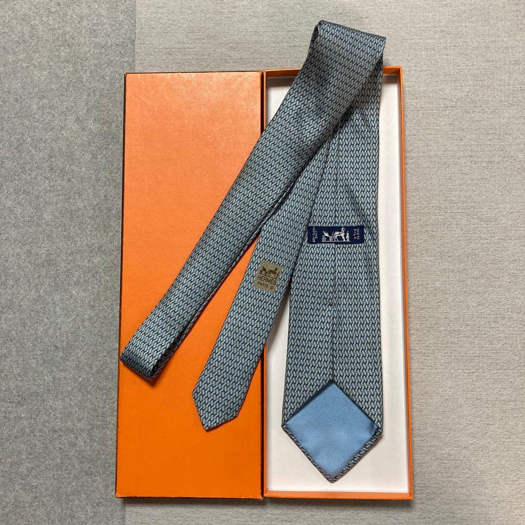 Blue-brown silk necktie with an H pattern laid inside an orange Hermès box showing the tie's back label and tip.