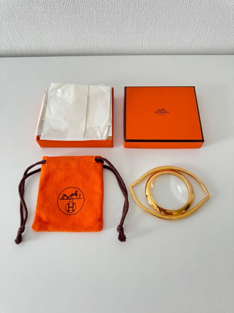 Gold-tone HERMES loupe shaped like an eye shown with its orange box and drawstring pouch.