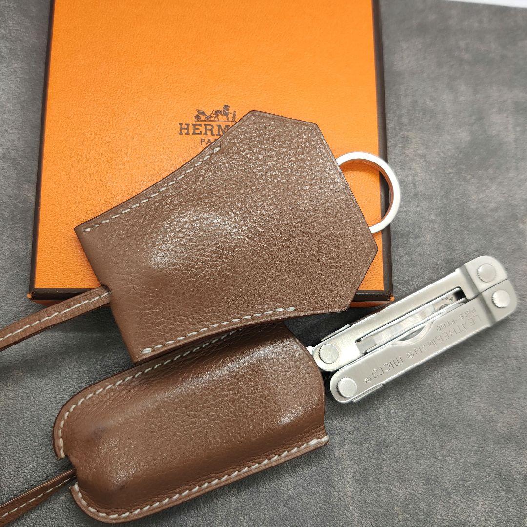 Brown Hermès leather clochette key holder on an orange Hermès box with a metal pocket tool beside it.